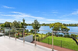 A balcony with a view of a lake, surrounded by greenery and a clear sky.