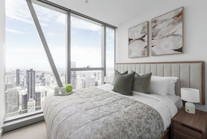 A bedroom with a large window, a bed with a grey floral bedspread, two pillows, and a nightstand with a lamp. - Sky High 2BR With Spectacular City Views (Melbourne)