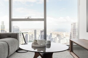 A modern living room with a large window, a round table with a tray of white cups, and a grey sofa. - Sky High 2BR With Spectacular City Views (Melbourne)