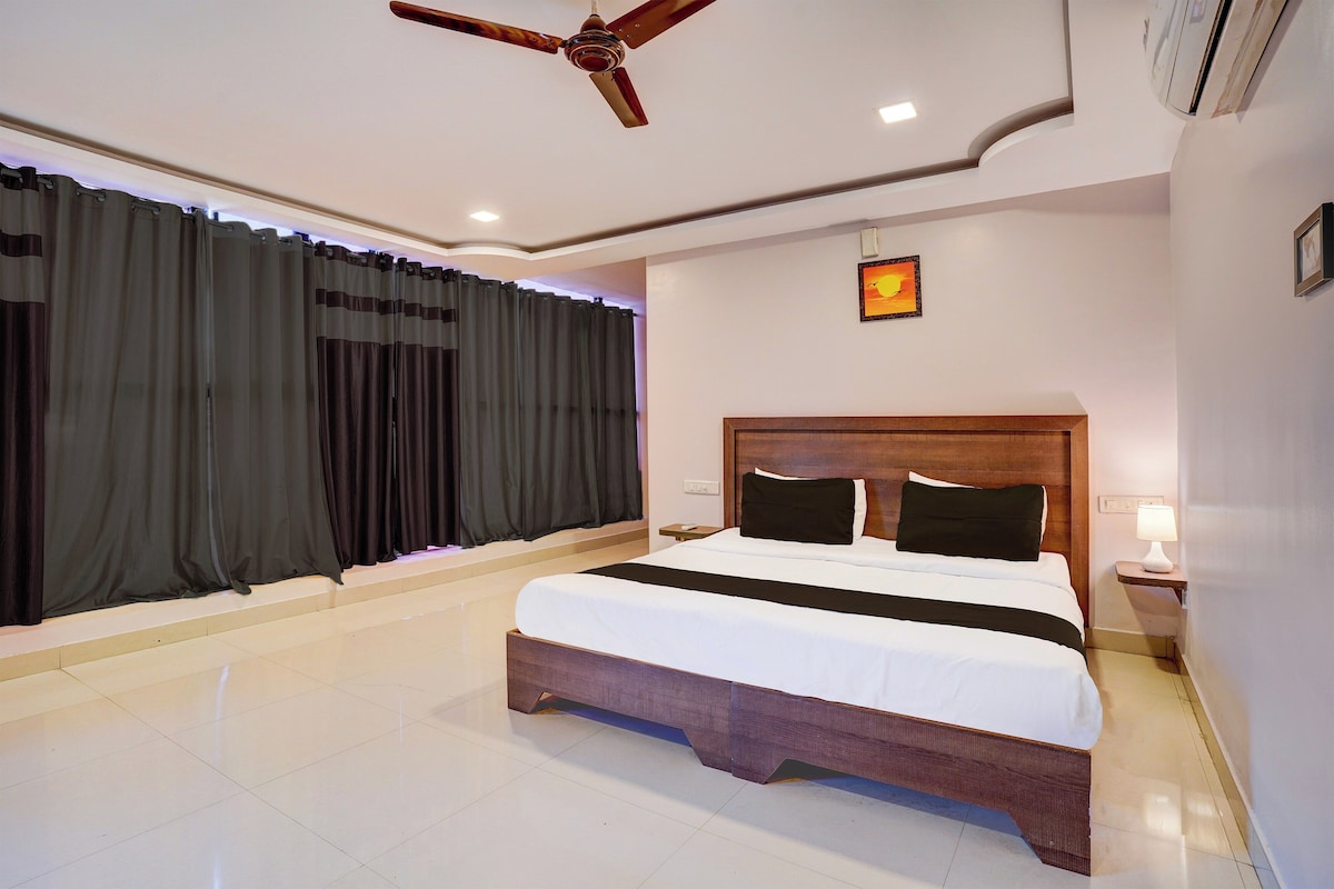 A hotel room with a bed, curtains, a ceiling fan, a nightstand with a lamp, and a wall-mounted picture.