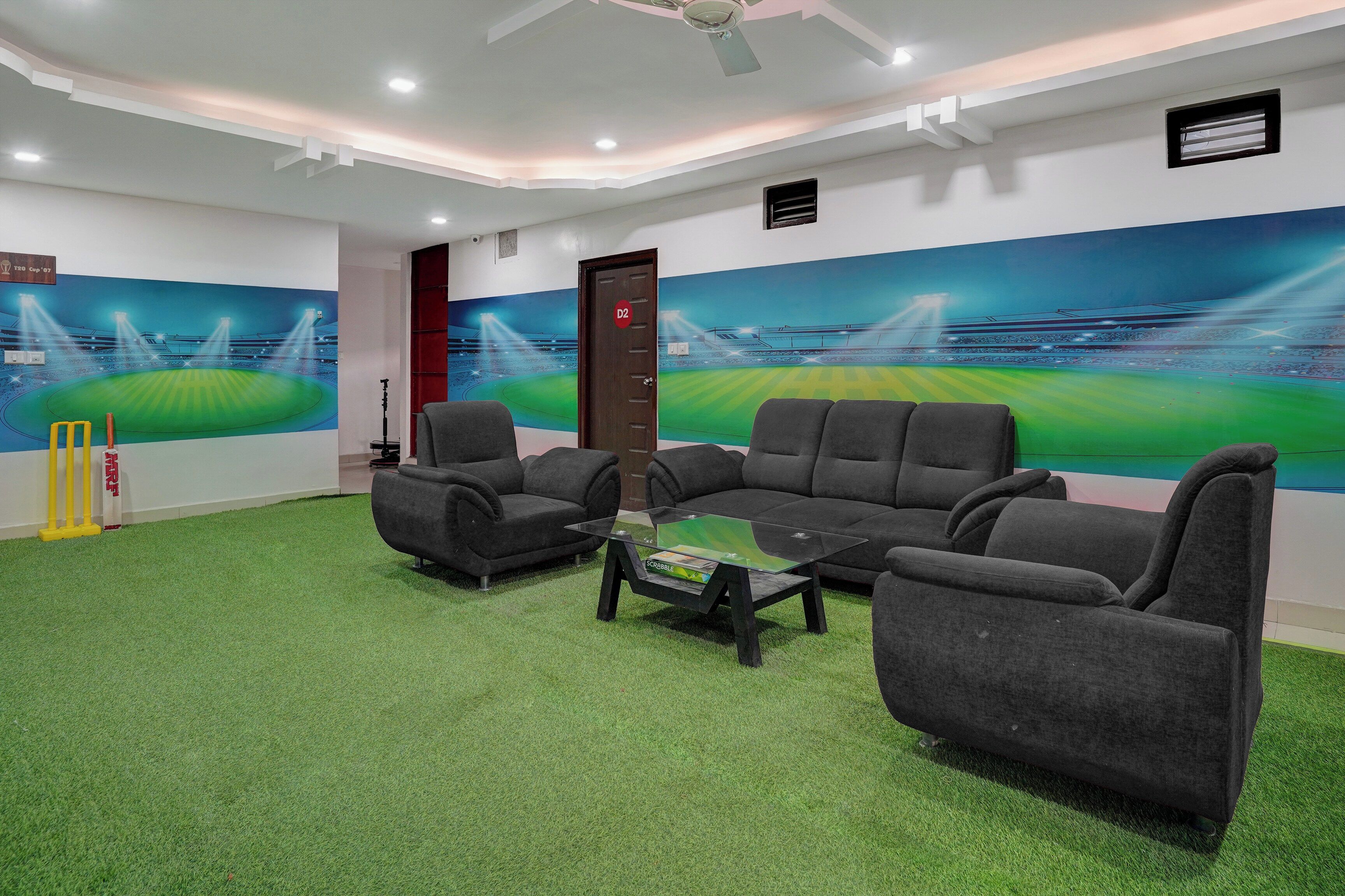 Una habitación con alfombra verde, dos sofás grises, una mesa de centro de vidrio y decoración de pared con temática de cricket.