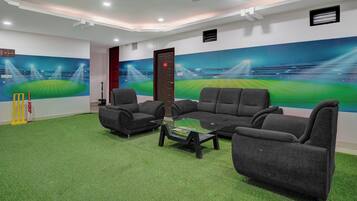Una habitación con alfombra verde, dos sofás grises, una mesa de centro de vidrio y decoración de pared con temática de cricket.