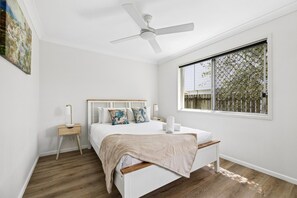 A bedroom with a bed, a nightstand, a lamp, a ceiling fan, and a window with a view of trees. - Ingara @ Maroochydore (Maroochydore)