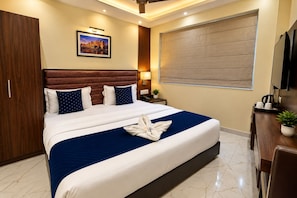 Habitación de hotel con una cama grande, cabecera de madera, mesita de noche, televisor montado en la pared y una ventana con persianas.