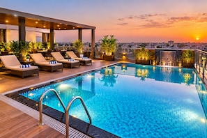 Una piscina infinita en la azotea con sillones de descanso y plantas en macetas, con vistas a un paisaje urbano al atardecer.