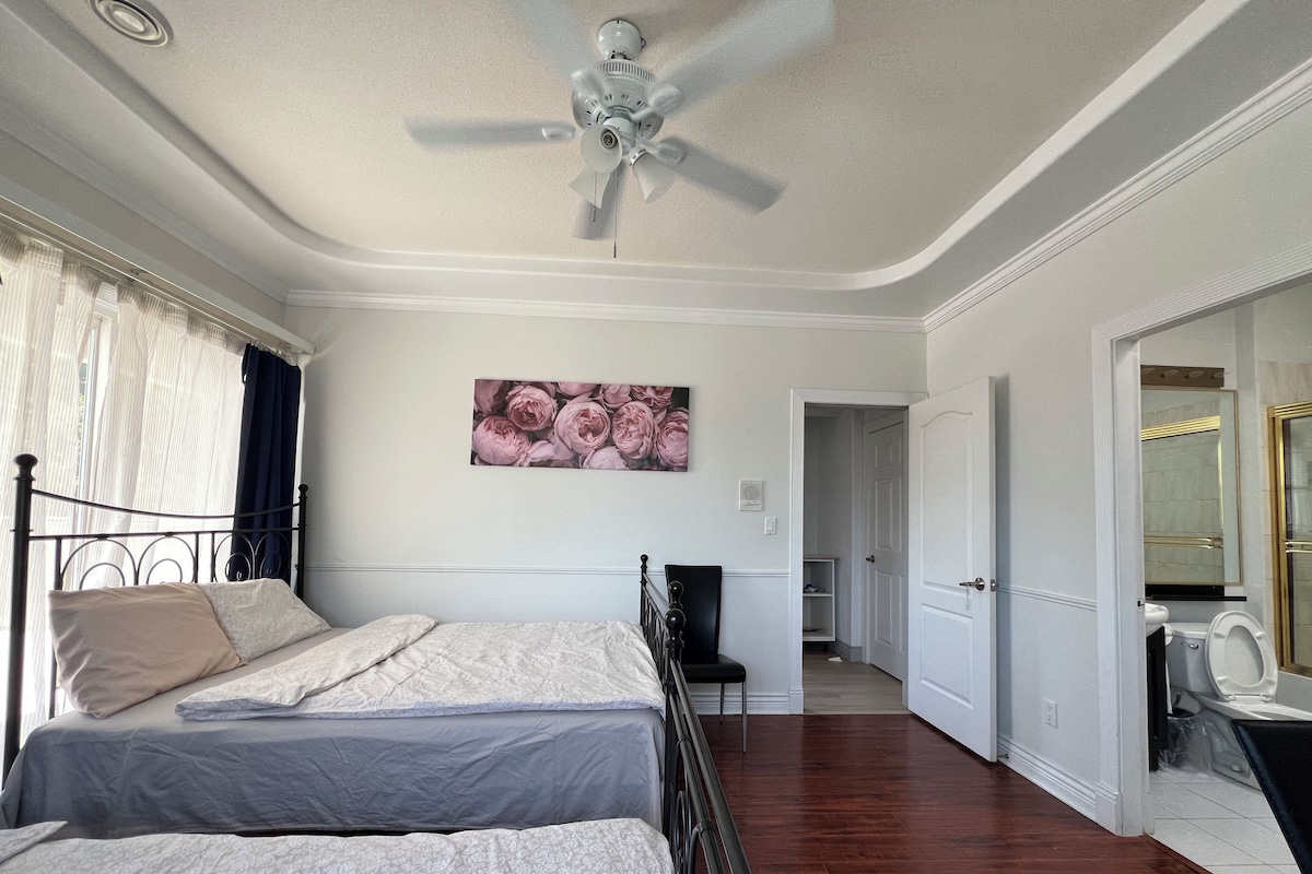 A bedroom with two beds, a ceiling fan, a painting of flowers, and a view of a bathroom.