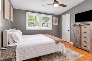 A bedroom with a bed, a ceiling fan, a window with blinds, a TV, and a wooden cabinet. - Midtown Executive | Work & Extended Stays | 4 Beds (Montgomery)