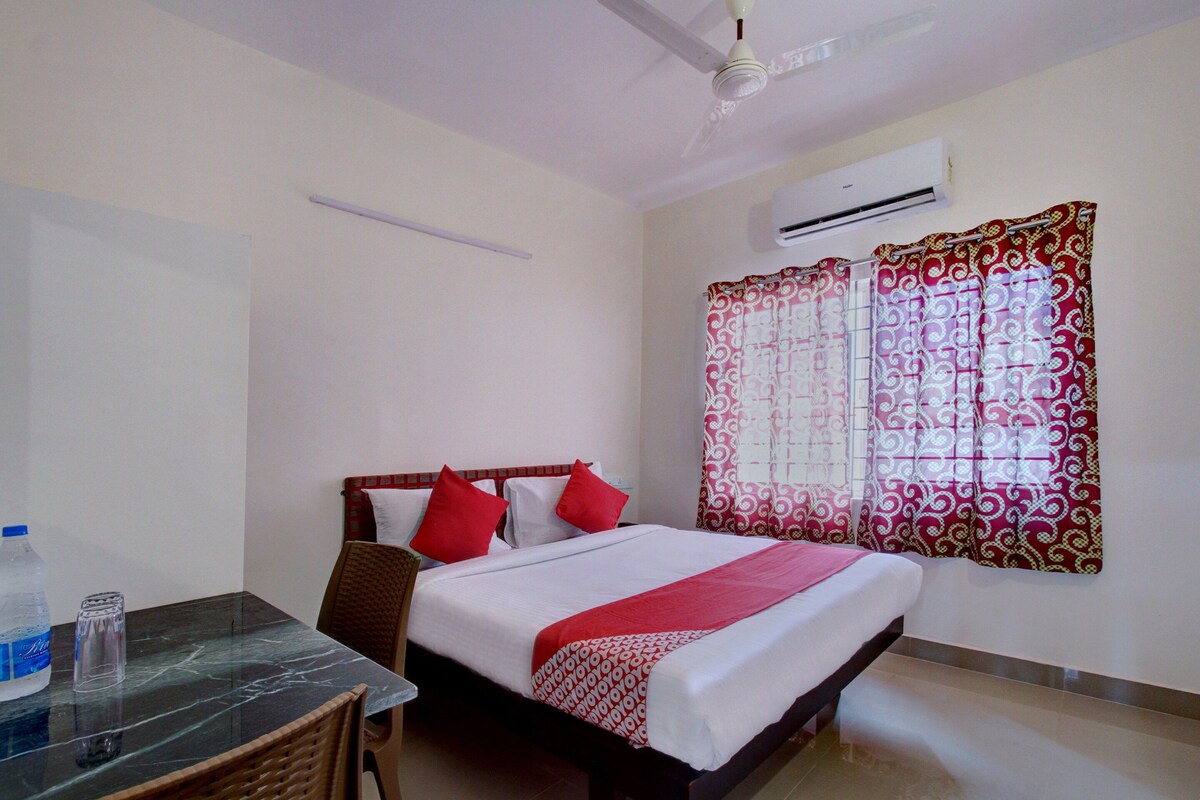 A hotel room with a bed, a table with chairs, an air conditioning unit, and a window with patterned curtains.