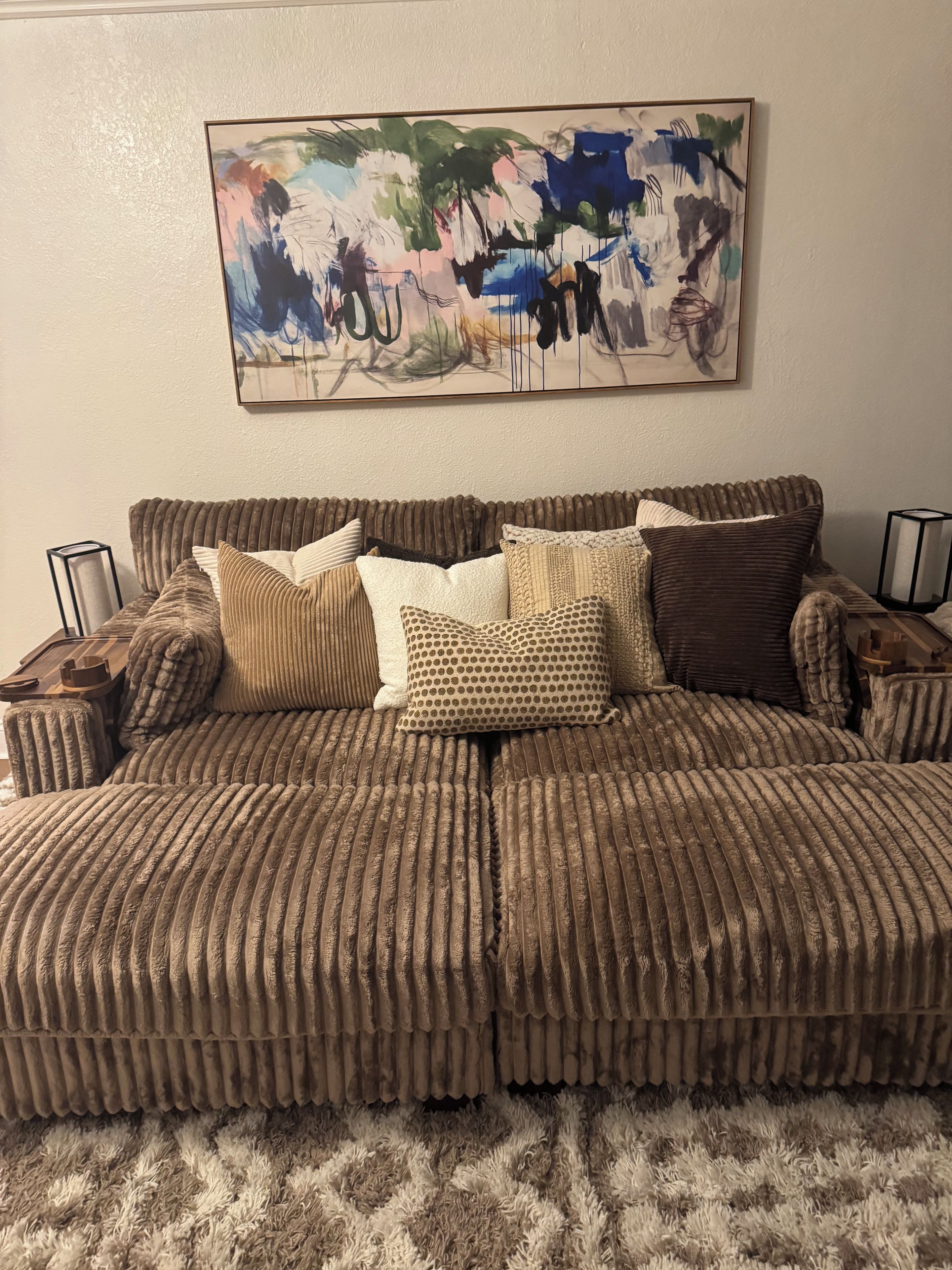 A sectional sofa with a textured fabric, multiple pillows, a framed abstract painting, and a fluffy rug.