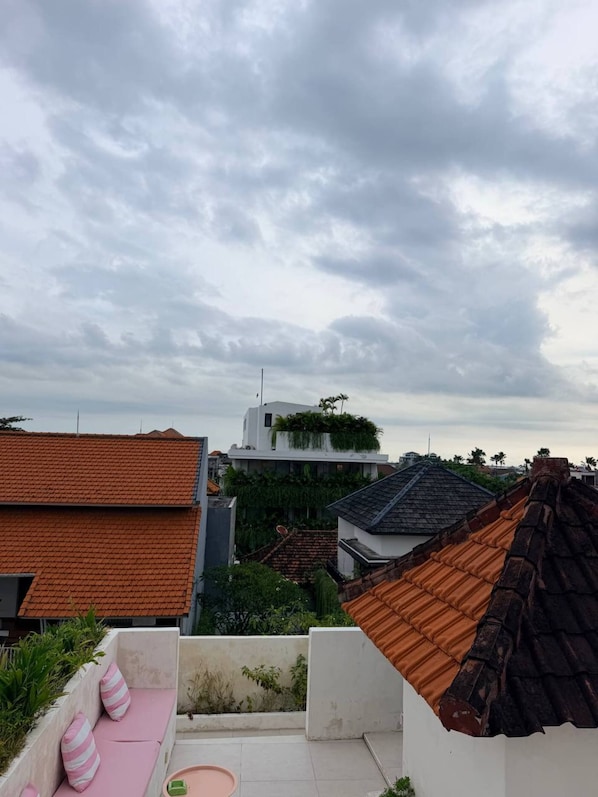 Eine Dachterrasse mit einer Sitzgelegenheit aus rosa Kissen, einem kleinen runden Tisch und Blick auf umliegende Gebäude.
