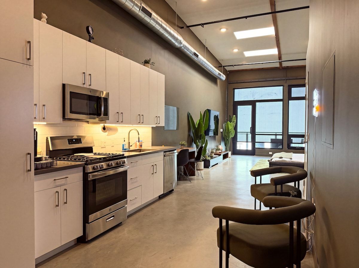 A modern kitchen with stainless steel appliances, a dining area with bar stools, and large windows providing natural light.
