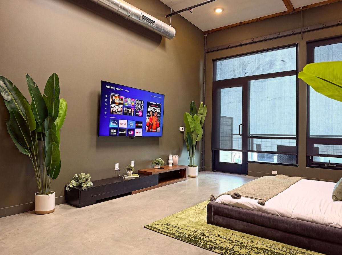 Una habitación de hotel moderna con un televisor grande montado en la pared, una cama con un tapete verde y ventanas amplias que dejan entrar luz natural.