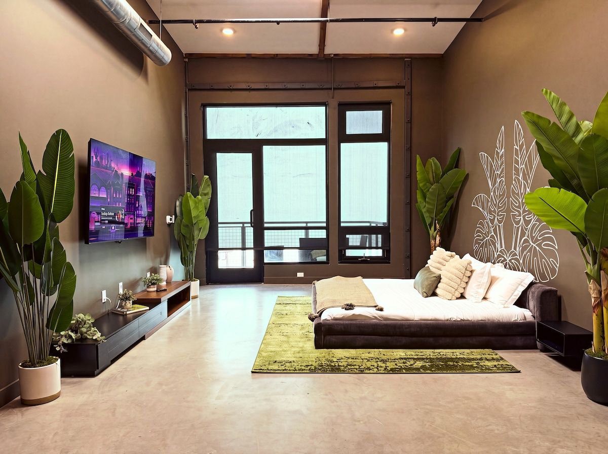 A modern living room with a large bed, a flat-screen TV mounted on the wall, and large windows.