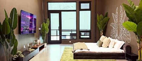 A modern living room with a large bed, a flat-screen TV mounted on the wall, and large windows.