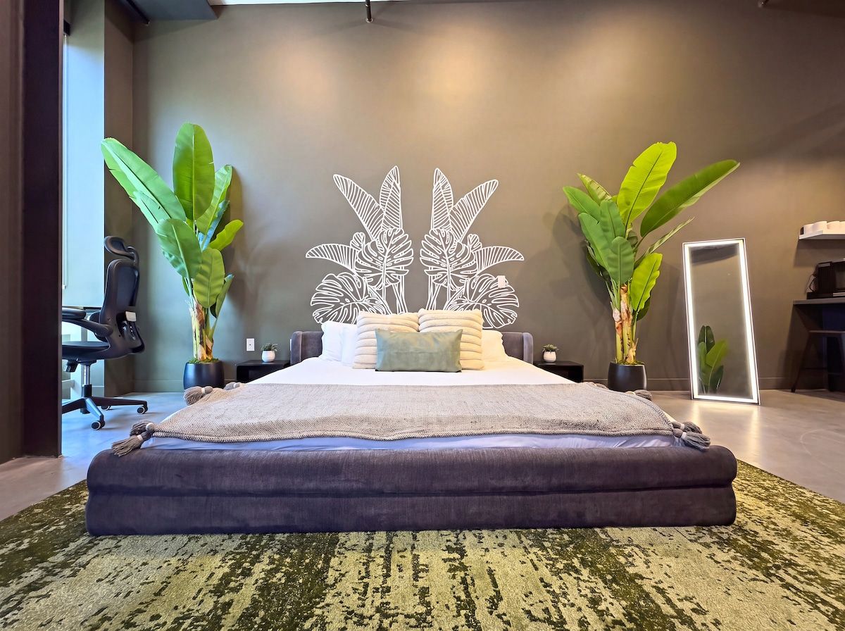 A modern bedroom with a bed, a large mirror, and two potted plants.
