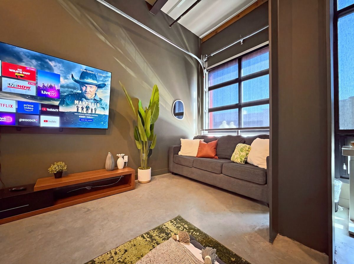A modern living room with a large flat-screen TV mounted on the wall, a grey sofa with cushions, a wooden entertainment unit, a potted plant, and a large window providing natural light.