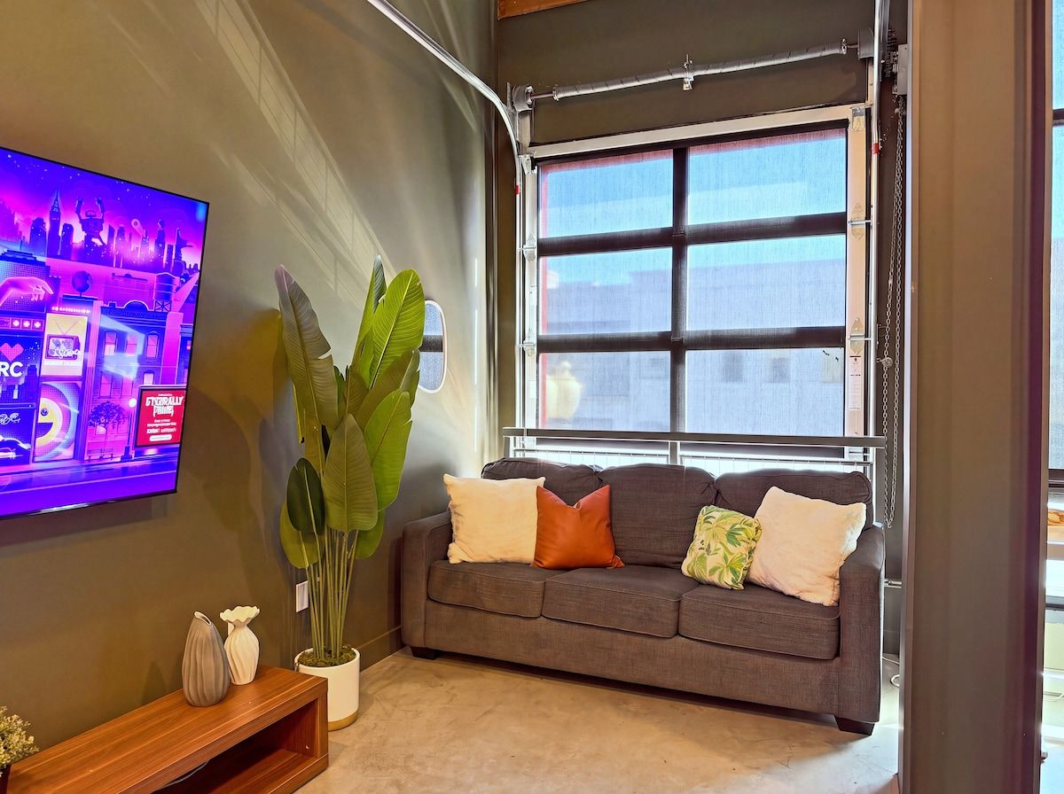 A living room with a grey sofa, a large potted plant, and a flat-screen TV mounted on the wall.