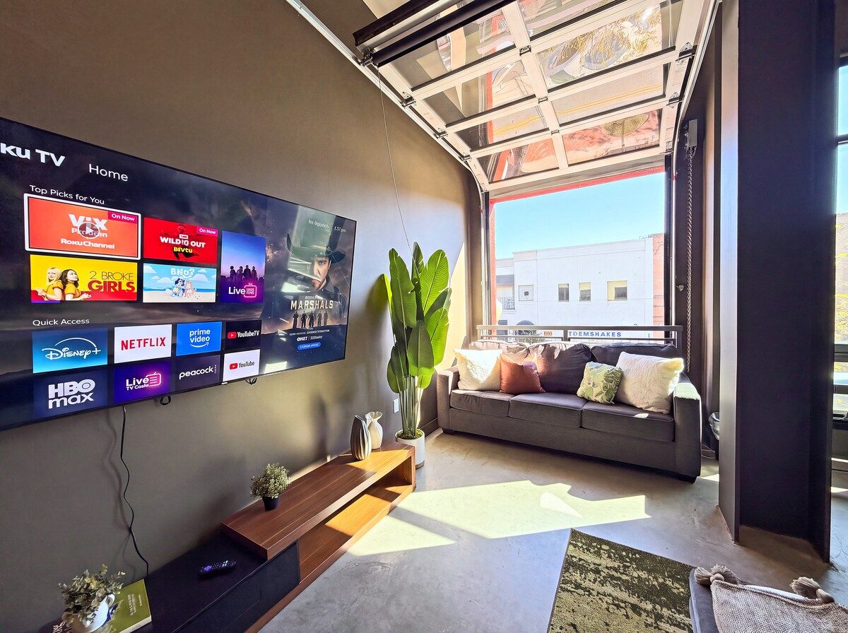 A modern living room with a large flat-screen TV displaying various streaming services, a grey sofa with cushions, a wooden side table, and a large window providing natural light.