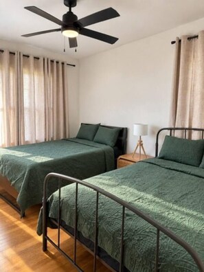 Ein Zimmer mit zwei Betten, einem Deckenventilator, einem Nachttisch mit Lampe und einem Vorhangfenster.