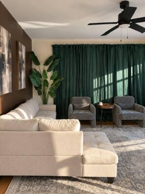 Un salon avec un canapé beige, deux fauteuils, une table basse, une grande plante en pot et un ventilateur de plafond.