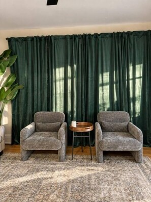 Ein Zimmer mit zwei Sesseln, einem kleinen runden Tisch und einem großen, grünen Vorhang.
