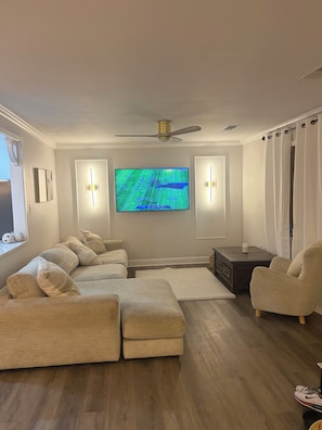 Uma sala de estar com sofá seccional, televisão fixada na parede, ventilador de teto e janela com cortinas.