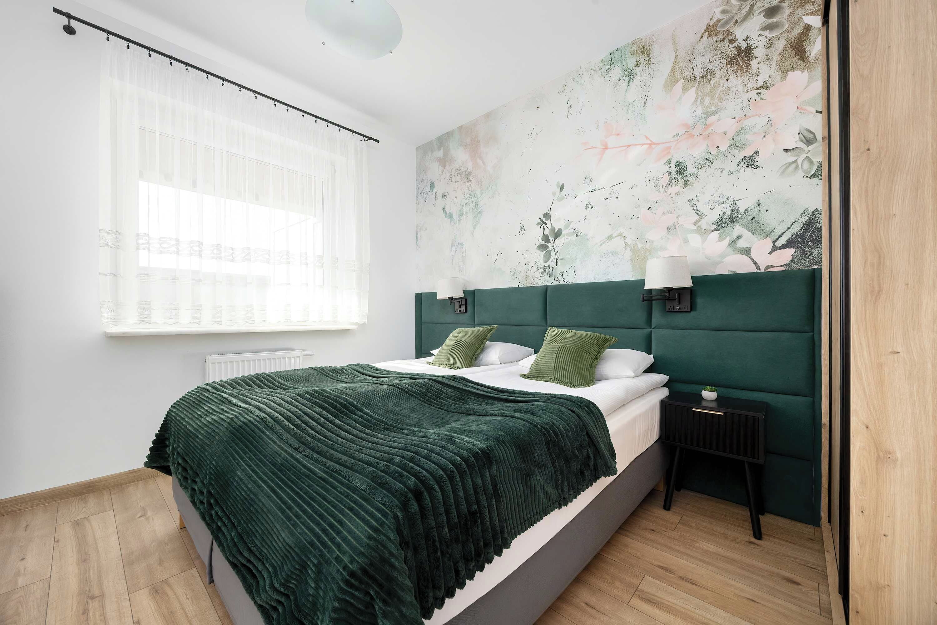 Ein Schlafzimmer mit einem Bett, einem Nachttisch, einer Wandlampe und einem Fenster mit durchsichtigen Vorhängen.