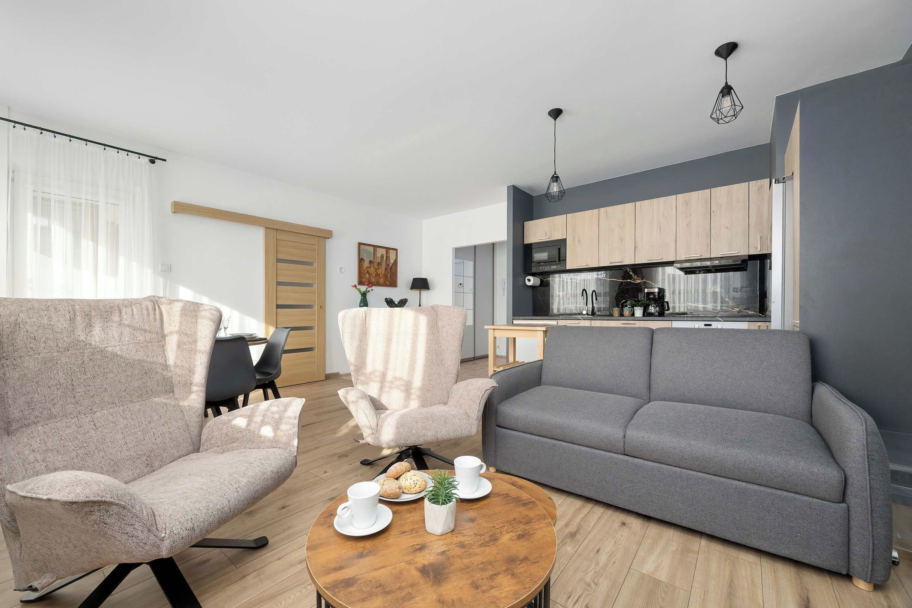 A modern living room with a grey sofa, a beige armchair, and a wooden coffee table. There is a kitchen in the background with wooden cabinets and a black countertop.