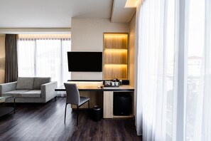 Ein modernes Hotelzimmer mit einer Couch, einem Schreibtisch mit Kaffeemaschine, einem Fernseher und Blick auf die Stadt durch das Fenster.