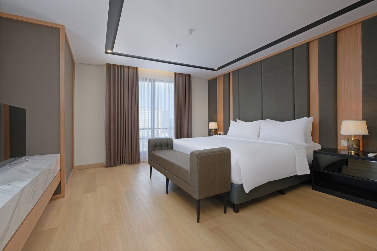 Una habitación de hotel con cama, mesitas de noche, escritorio, silla y un amplio ventanal con cortinas.