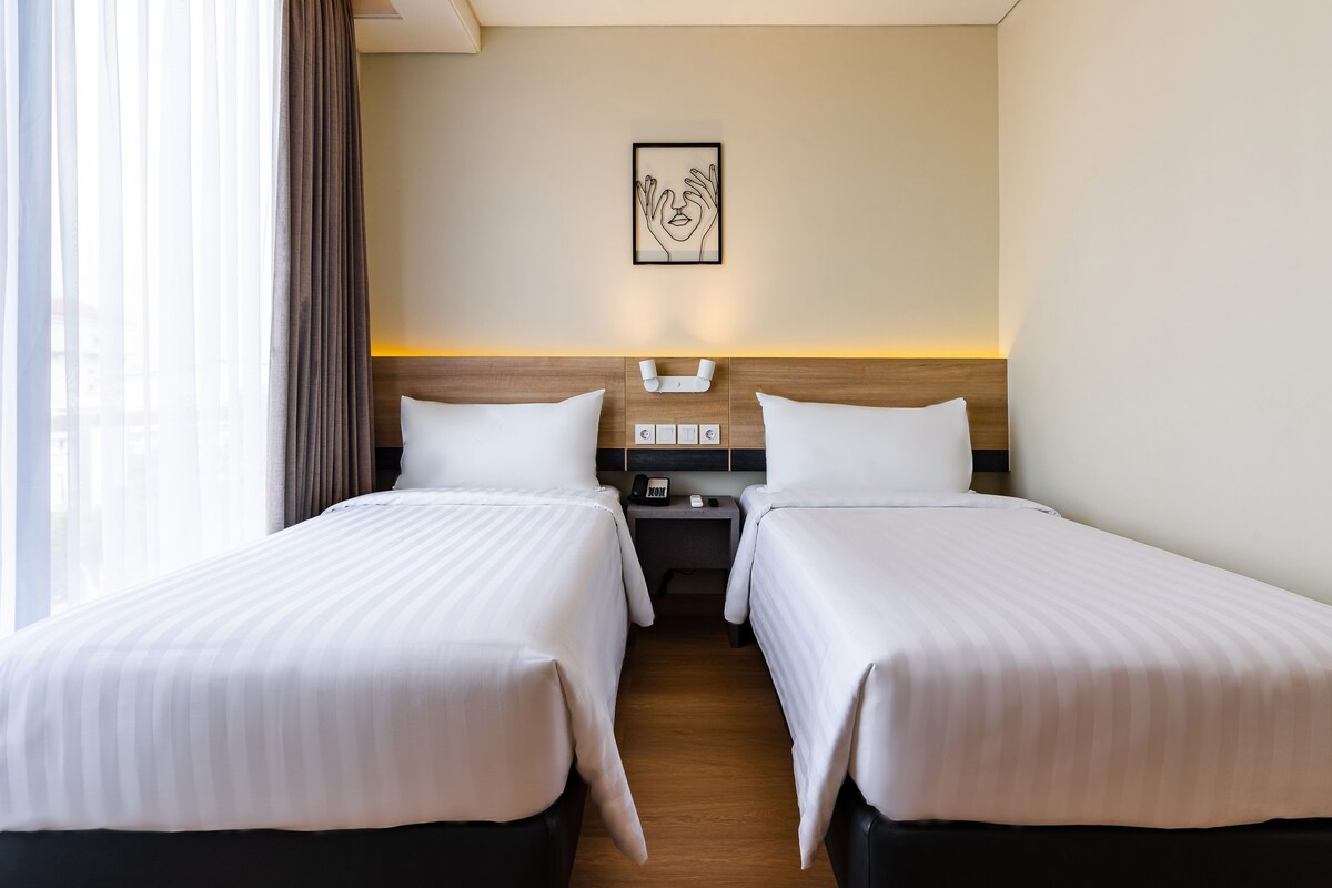 Una habitación de hotel con dos camas individuales, una mesita de noche con una lámpara, un teléfono en la pared y una obra de arte enmarcada sobre la cama.