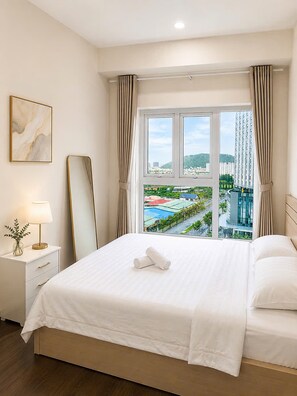 A hotel room with a large bed, a bedside table with a lamp, a mirror, and a view of a cityscape through a window. - Mermaid Seaview-Apartment near beach (Vũng Tàu)