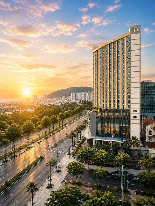 A modern hotel with a prominent glass facade, situated beside a wide, tree-lined avenue during sunset. - Mermaid Seaview-Apartment near beach (Vũng Tàu)