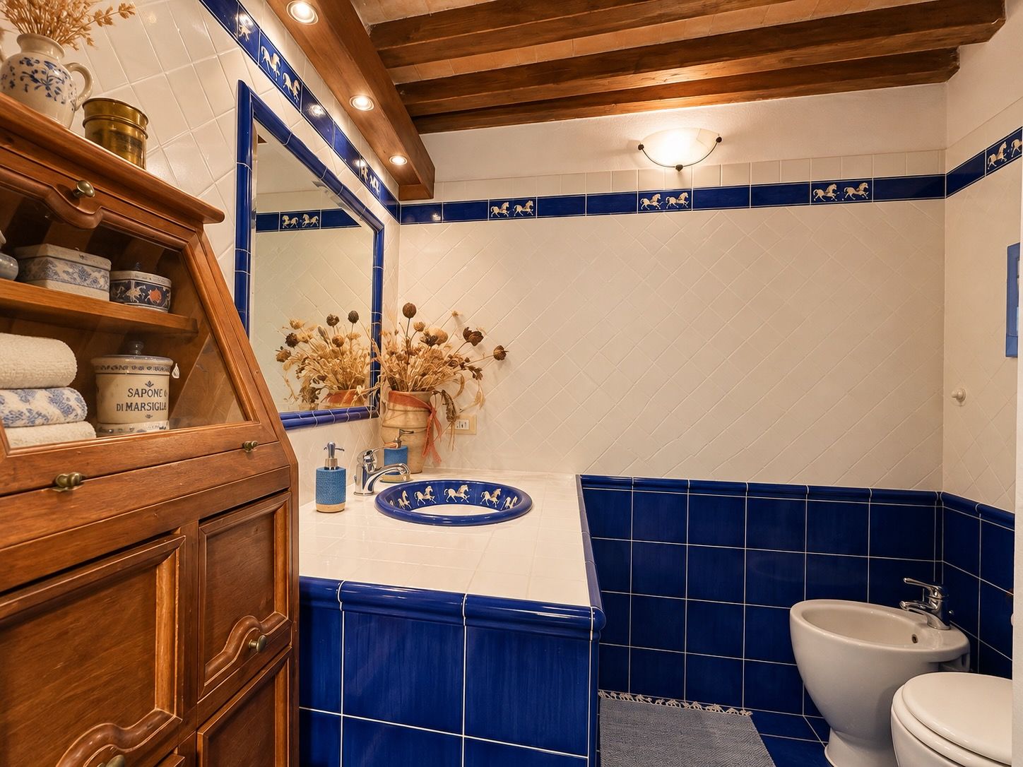A bathroom with a wooden cabinet, a mirror, a sink with blue tiles, a toilet, and a decorative vase with dried flowers.