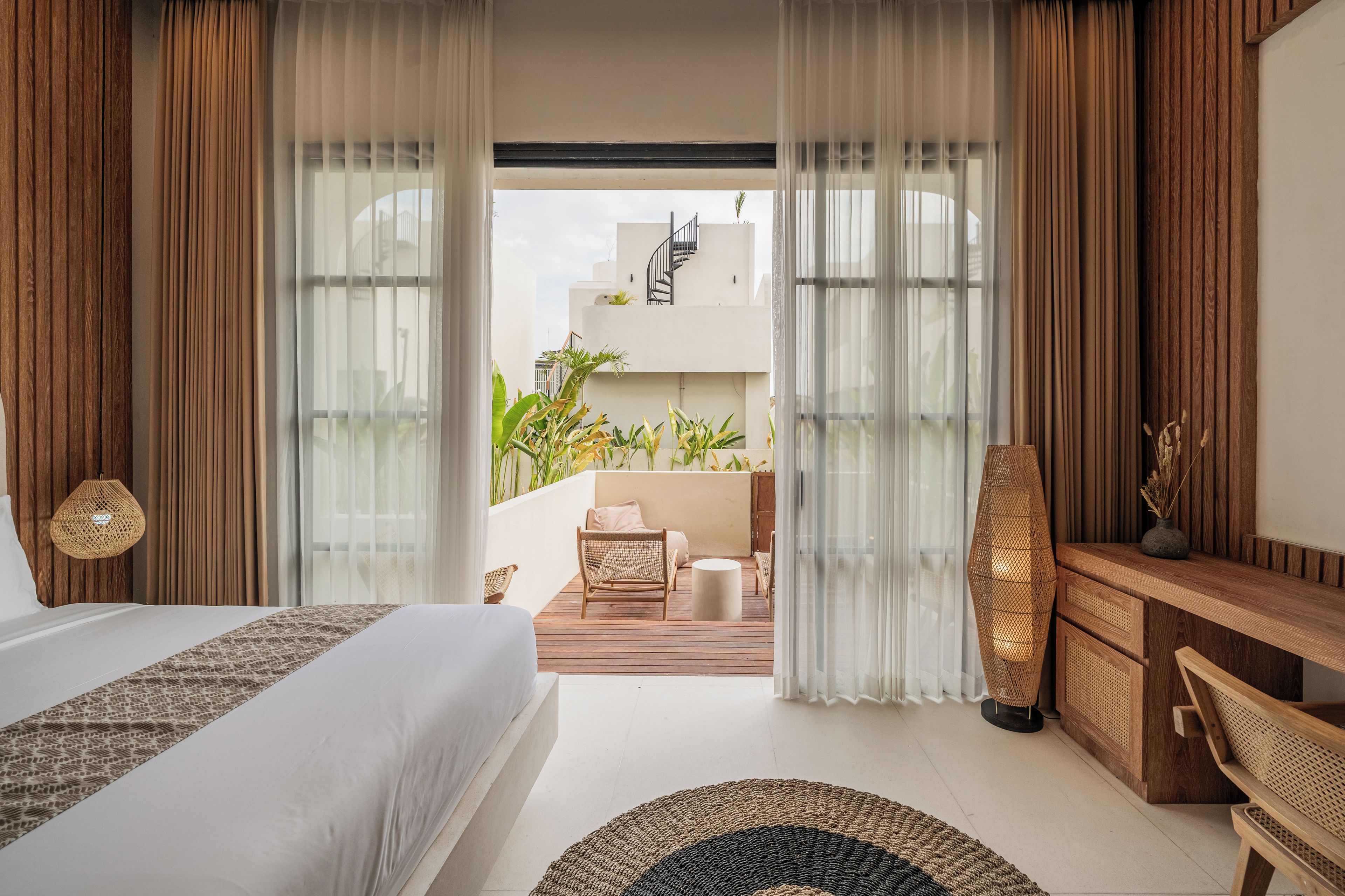Une chambre d’hôtel avec un lit, un bureau en bois, un tapis rond et un balcon agrémenté de plantes et d’une chaise.
