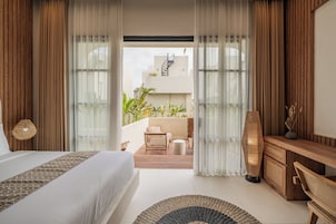 Habitación de hotel con cama, escritorio de madera, alfombra redonda y balcón con plantas y una silla.