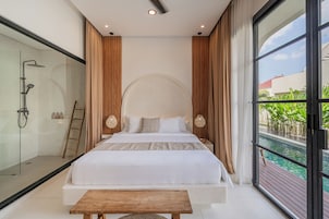 A modern bedroom with a large bed, a wooden bench, a glass shower enclosure, and a view of a swimming pool.