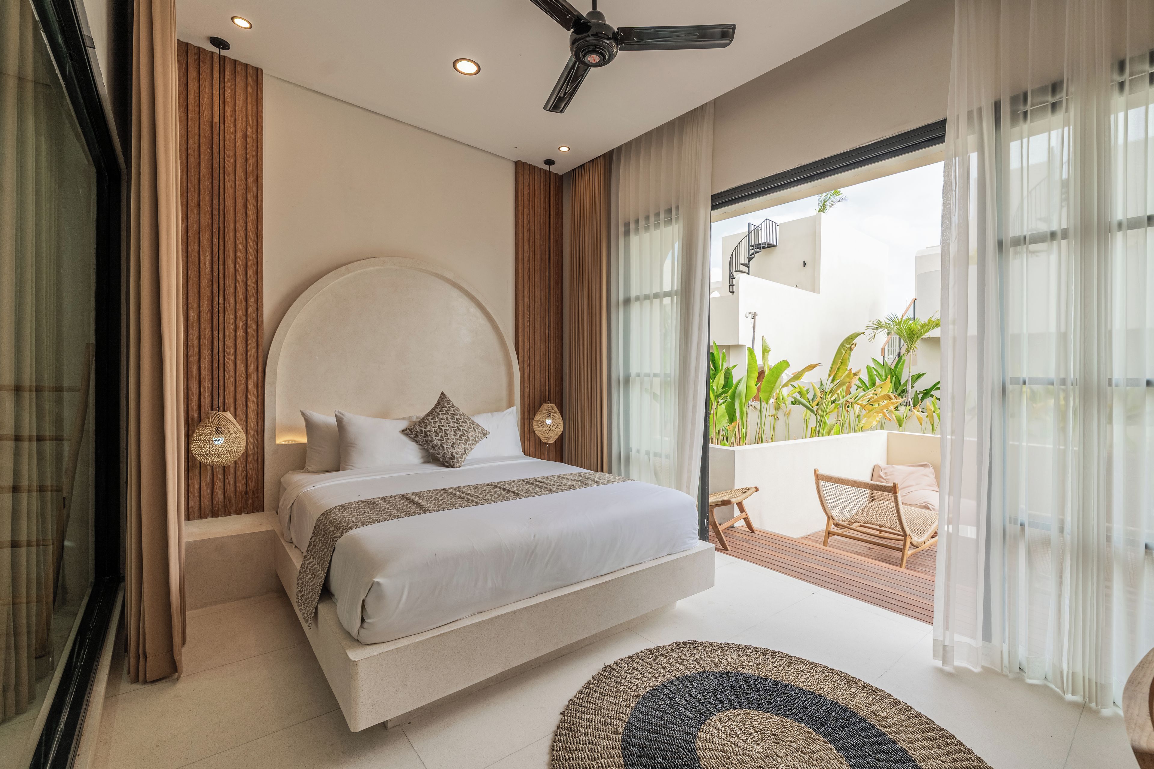 A modern bedroom with a large bed, a ceiling fan, and a view of a balcony with plants.
