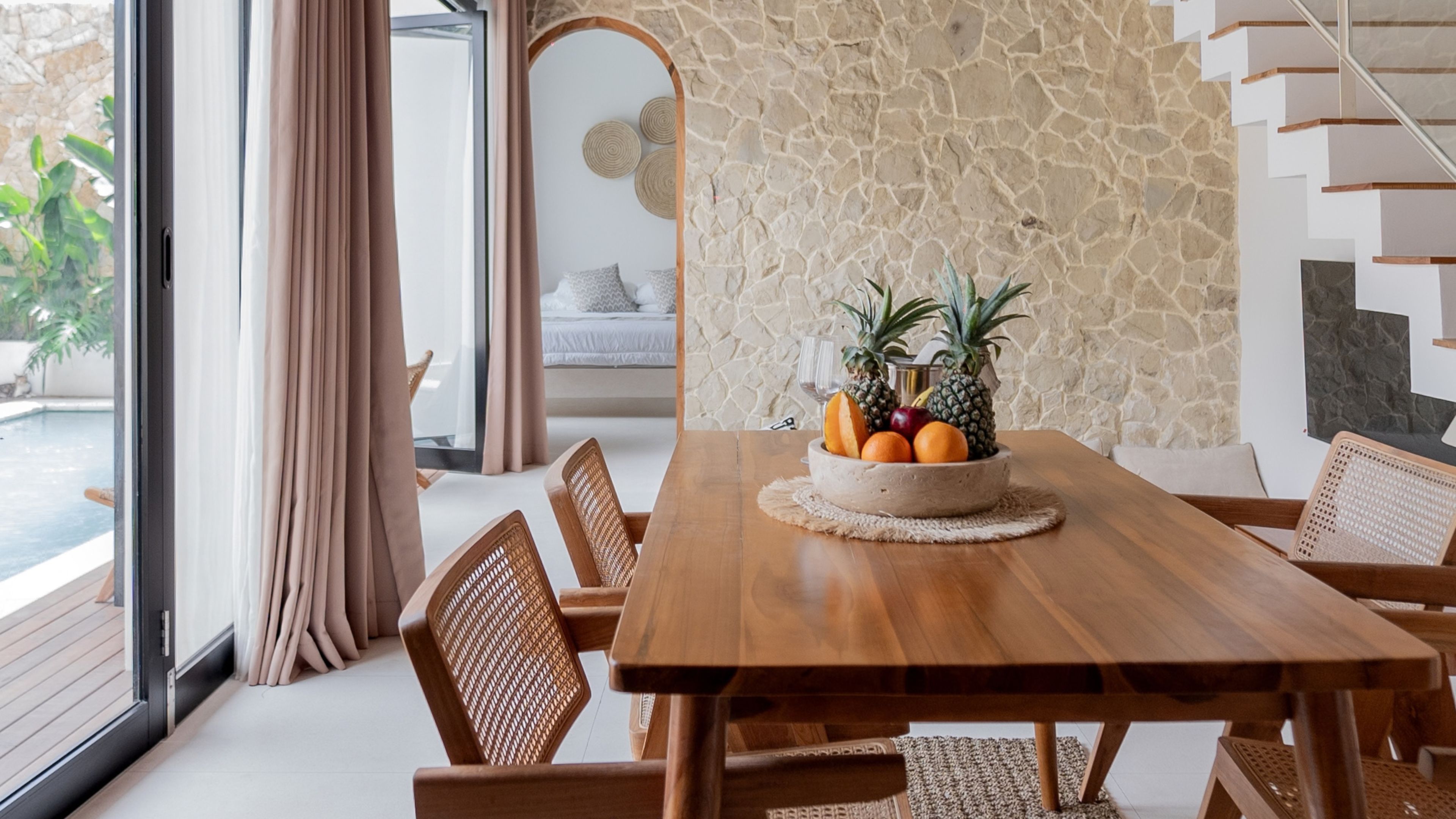 Una mesa de comedor de madera con un frutero en el centro, sillas de mimbre, un muro de piedra y vistas a una piscina a través de amplias ventanas.