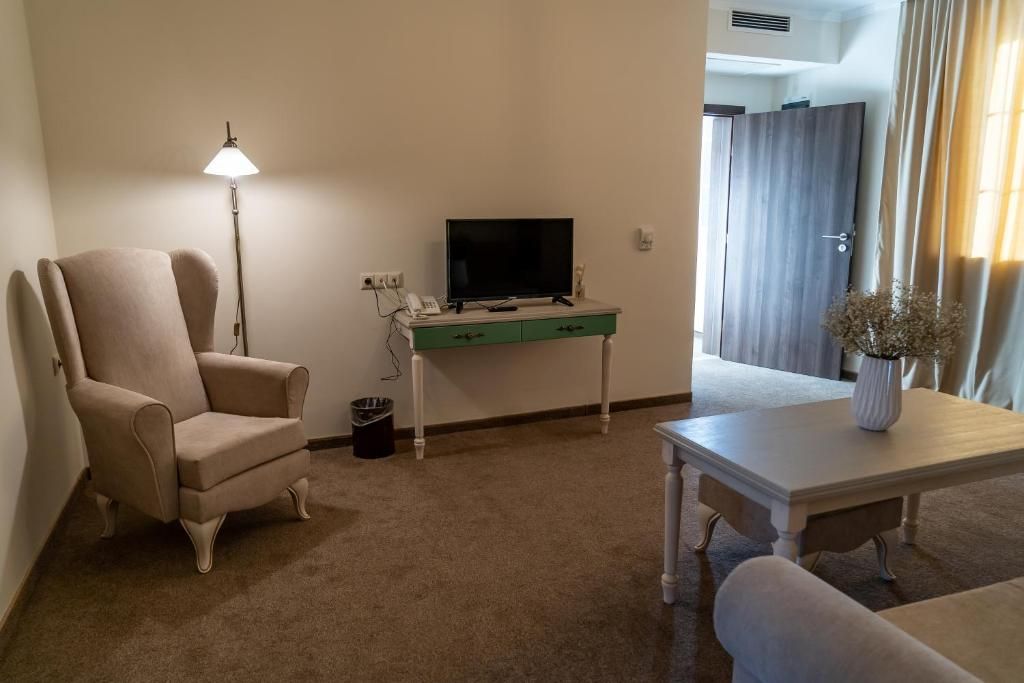 Una habitación con un sillón color beige, un televisor sobre un escritorio verde, una mesa blanca con un florero, y una puerta con cortinas.