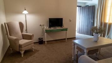 Una habitación con un sillón color beige, un televisor sobre un escritorio verde, una mesa blanca con un florero, y una puerta con cortinas.