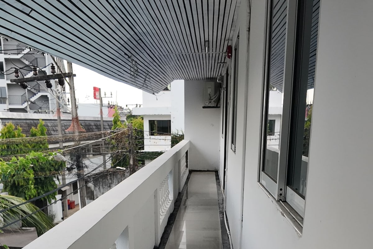 Un pasillo estrecho con barandilla blanca, suelo de baldosas y vistas a edificios y vegetación al exterior.