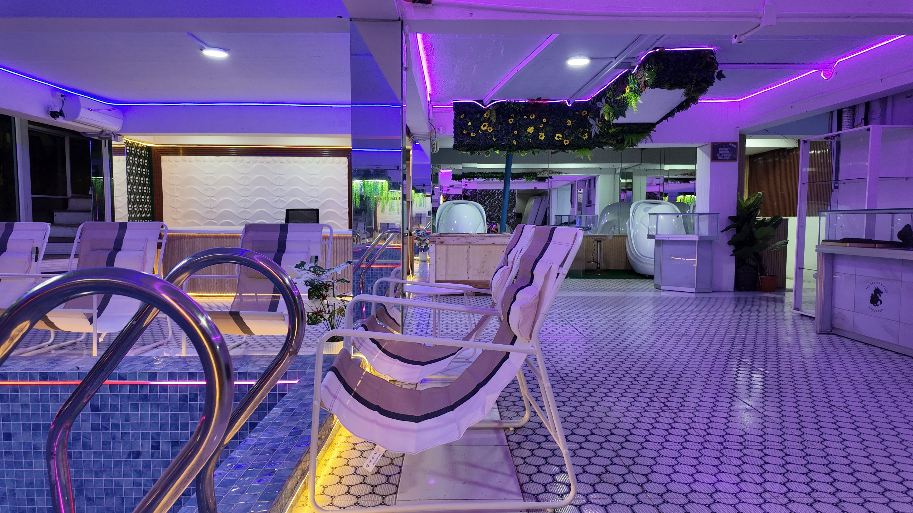 A modern indoor pool area with lounge chairs, a glass balustrade, and a ceiling with purple LED lighting.