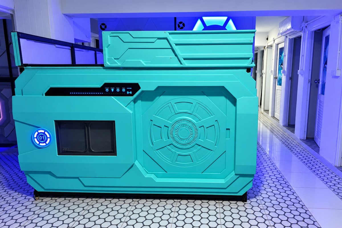 Un equipo de color aguamarina, de aspecto futurista, con un panel de control y un elemento de diseño circular, ubicado en un pasillo con suelo de baldosas y puertas.