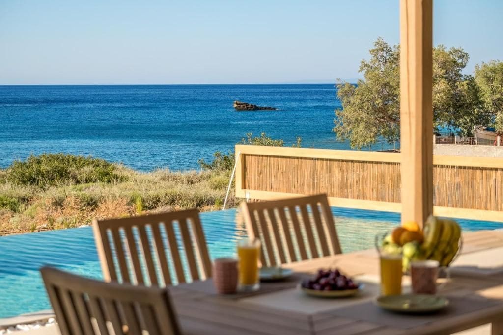 Mesa con sillas, frutero y vaso de jugo, con vista a una piscina y al mar.