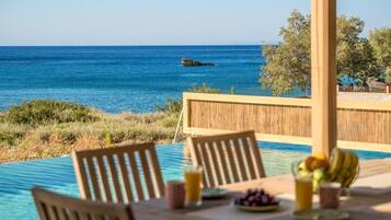 Mesa con sillas, frutero y vaso de jugo, con vista a una piscina y al mar.