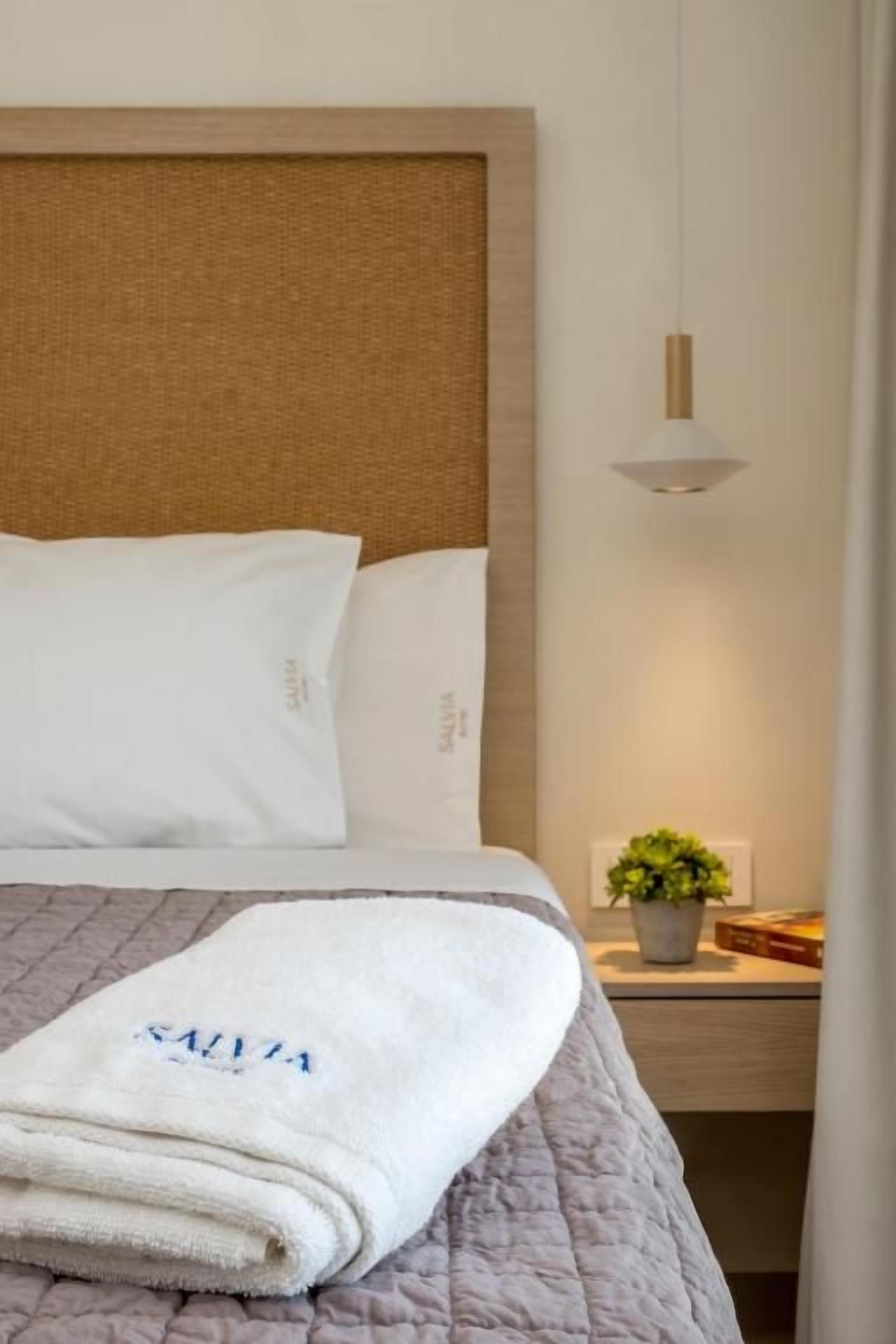 A neatly made bed with a white blanket and a towel with text, a bedside table with a plant, and a hanging light fixture.