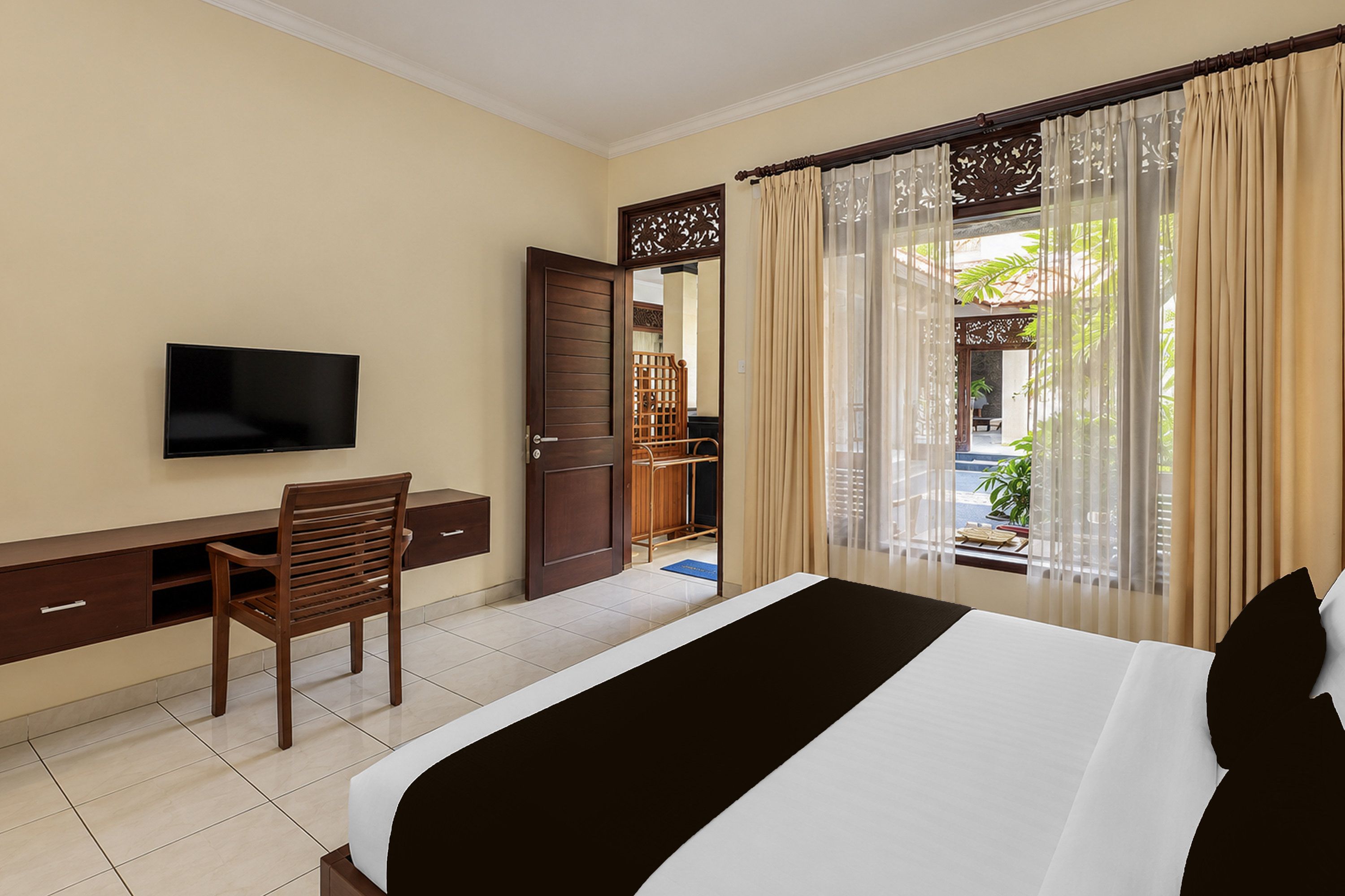 A hotel room with a bed, a desk, a chair, a television, and a view of a garden through a window with curtains.