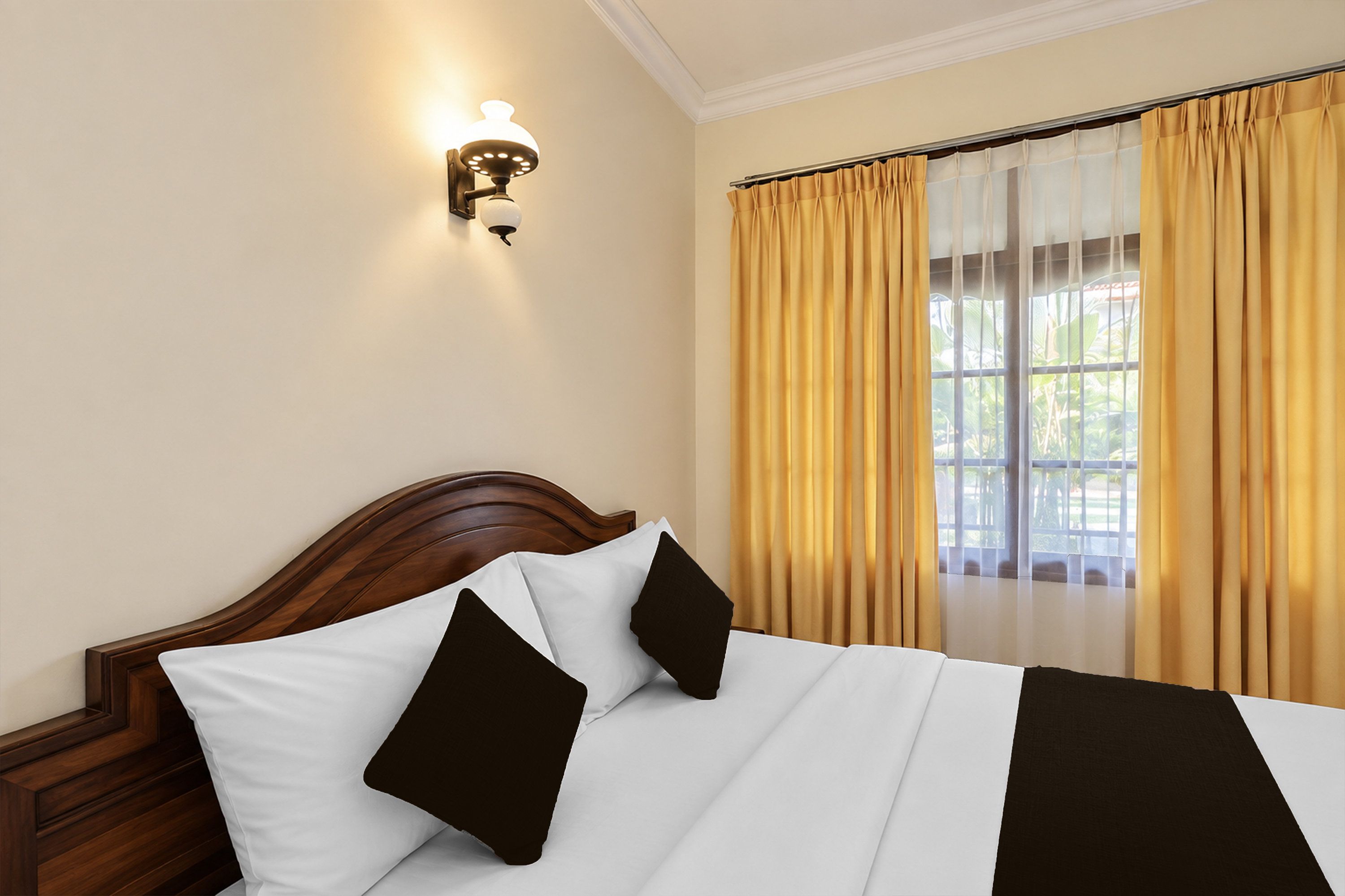 Habitación de hotel con cabecera de madera, cama con ropa de cama blanco y negro, lámpara de pared y ventana con cortinas amarillas.