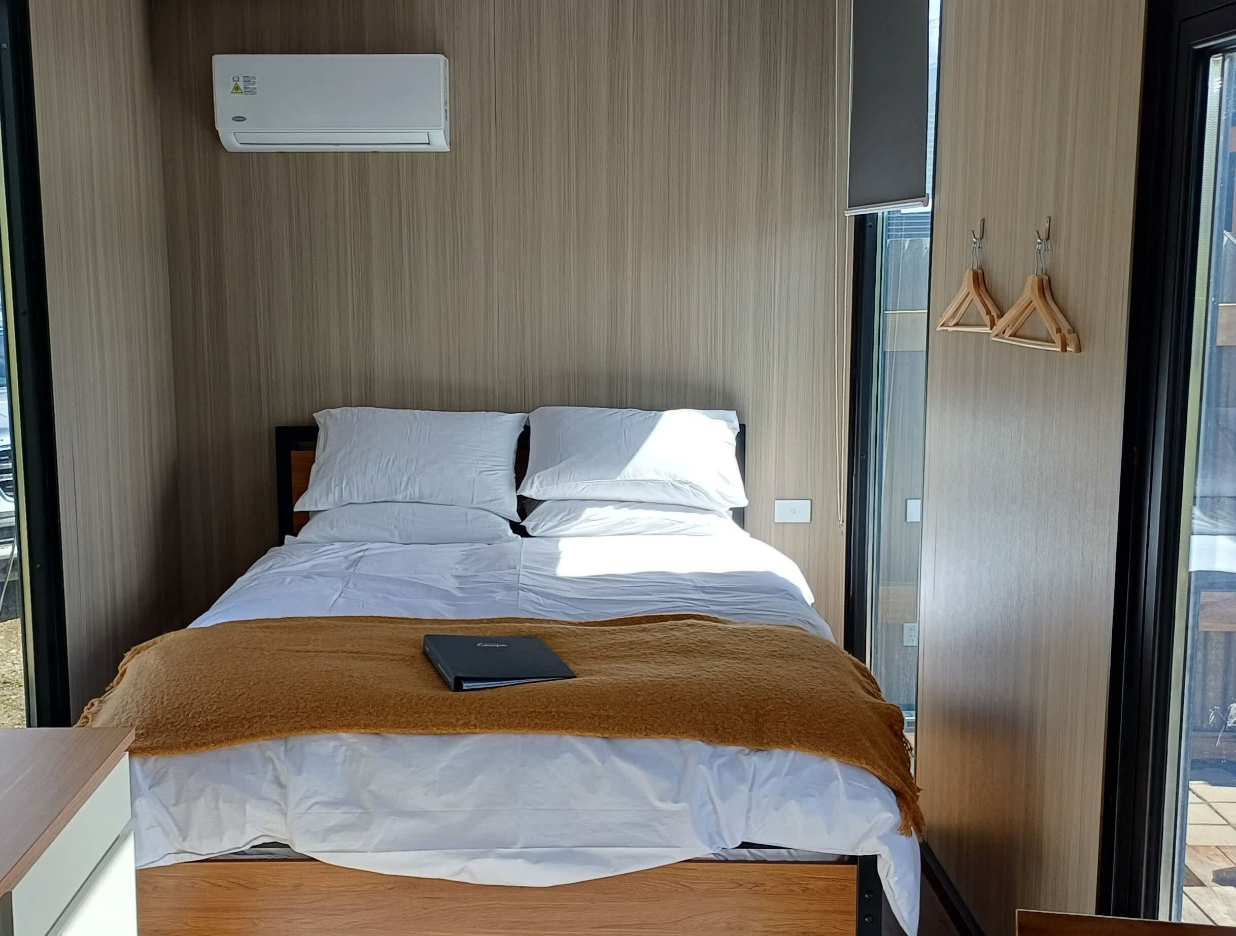 A modern hotel room with a bed, a brown throw blanket, a bedside table, a window, and an air conditioning unit.